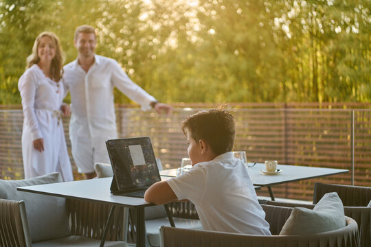 Family On Balcony With Parents Watching Son Use Laptop At Sunset On Modern Terrace
