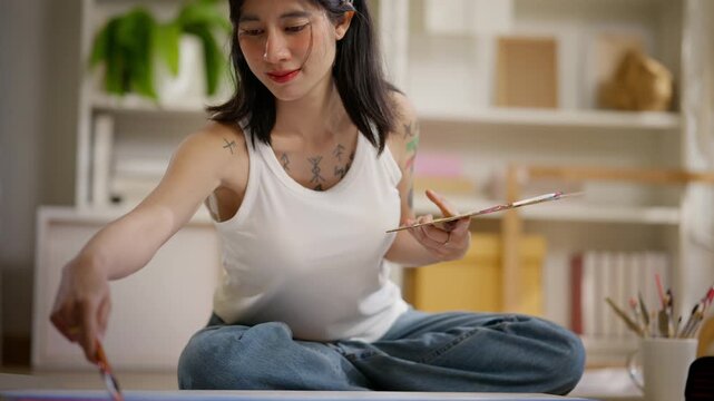 Young Asian female artist sitting on the floor, holding a palette and painting on a canvas framework. Creative girl enjoying painting as an art therapy hobby. lifestyle and home activity.