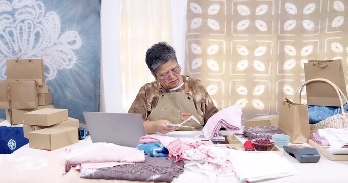 Senior asian woman entrepreneur studies invoice list, matches receipt total with online order record on laptop, sorts tie-dye shibori fabric, prepares packing plan for parcel shipping at home studio.