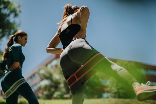 Two women perform a resistance band leg exercise outdoors in a park. The pair train with loop bands during a sunny outdoor fitness session.