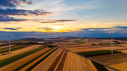 Obraz premium Wind turbines over rural fields at sunset. Drone view with village, hills, golden crops, layered clouds.