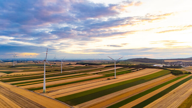 Wind turbines stand tall in green and yellow fields during sunset. Wind turbines spin in a landscape of fields at sunset. The sky is bright with colors.