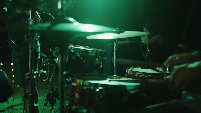A close-up of a drummer playing a drum kit under dramatic green stage lighting, focusing on the metallic cymbals and snare drum during a live music performance or concert.