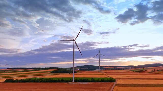Wind turbines standing in cultivated fields near distant hills. Panoramic view of alternative power generators during bright sunset with yellow crops and green trees.