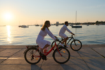 Fototapeta premium Couple Cycling Along Waterfront At Sunset Near Sailboats On Summer Evening