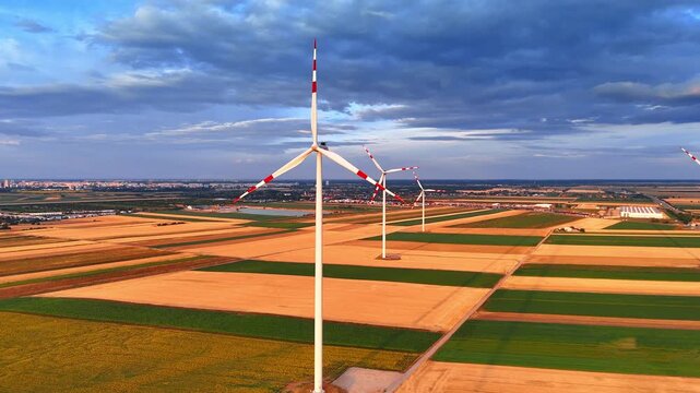 Wind turbines standing in agricultural fields under sunset sky. Aerial view of renewable energy generators among colorful crop plots with distant city building silhouettes.