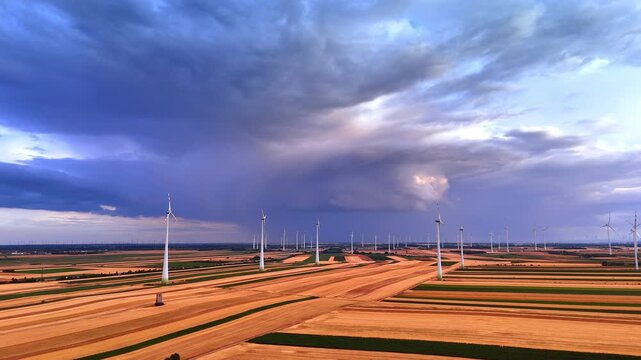 Multiple wind turbines operating across striped agricultural fields. Wide aerial perspective of clean energy farm under dramatic dark blue storm clouds and overcast sky.