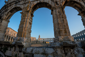 Obraz premium Pula, Croatia - August 22, 2025: View through ancient stone arches of a Roman amphitheater at sunset, revealing the inner arena and distant tower.