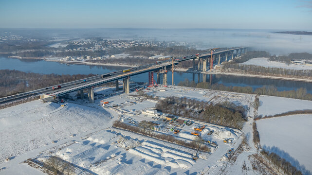 Highway bridge construction site over the Kiel Canal in winter landscape, with traffic moving on the existing lanes while large cranes and construction equipment work on building new bridge. 