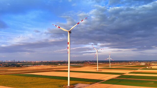Wind turbines working in agricultural fields under cloudy sky. High view of alternative power structures surrounded by yellow crops with city horizon under dark rain clouds.