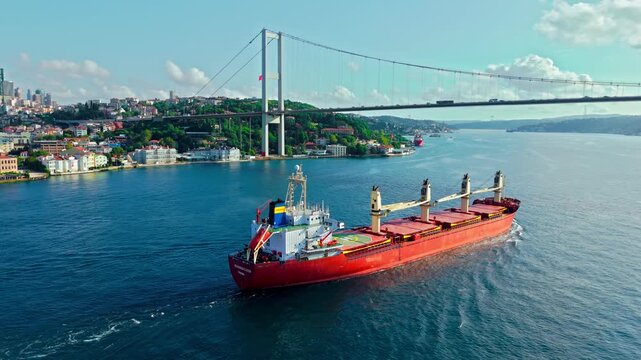 Drone view of a red cargo ship on the Bosphorus near the Bosphorus Bridge. The vessel moves across calm water with the bridge and Istanbul coastline in the background.