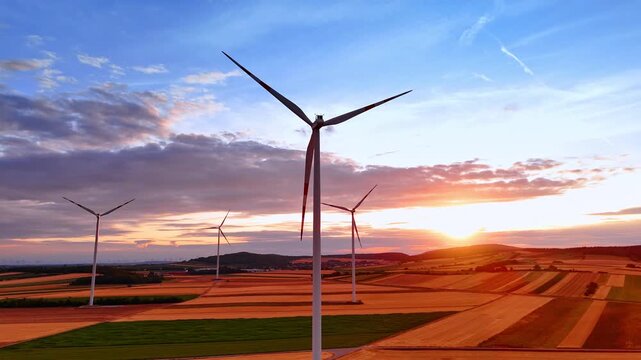 Sustainable wind turbine spinning at vibrant summer sunset. Low angle aerial shot of modern power generator over orange agricultural fields.