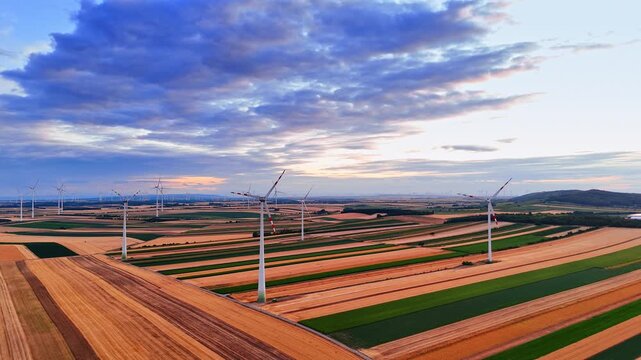 Clean energy wind farm situated in vast countryside. High angle drone view of eco turbines on multicolored fields under stormy sky.