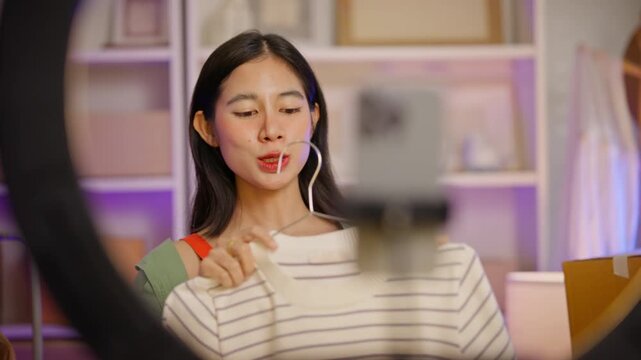 Cheerful young Asian woman merchant holding a shirt while live streaming to sell clothes via smartphone with a ring light. Female entrepreneur running an e-commerce fashion business from home.