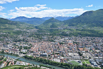 Fototapeta premium Panoramic aerial view of Trento, Italy, featuring the historic city center, the Adige River, green valleys, vineyards, and the snow-capped Alps under a blue summer sky. 