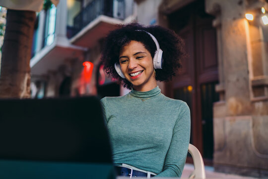 young Latin female laughing while watching streaming social media video options on portable tablet screen, happy Gen Z tourist using wireless internet connection on cozy coffee shop terrace