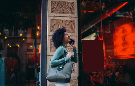 young student talking on a mobile phone while standing on the street near restaurant with bright red window lighting, happy Gen Z tourist carrying handbag during night walk conversation