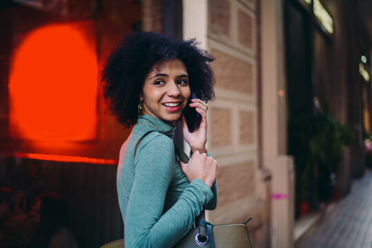 young woman turning her head while smiling during a cheerful cell phone call conversation near bright red street neon light, happy Gen Z mobile user talking with friends on a walk outside