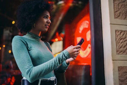 young person checking chat notifications on a mobile phone touchscreen display near bright red neon window light at night, focused Gen Z user waiting for message feedback on street walk