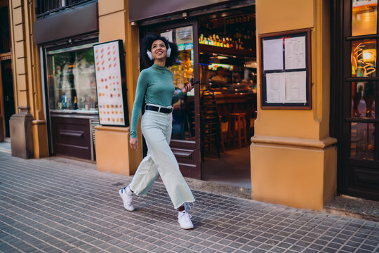 young student enjoying digital music streaming through wireless headphones while strolling along city street, happy Gen Z female tourist holding smartphone device near local restaurant
