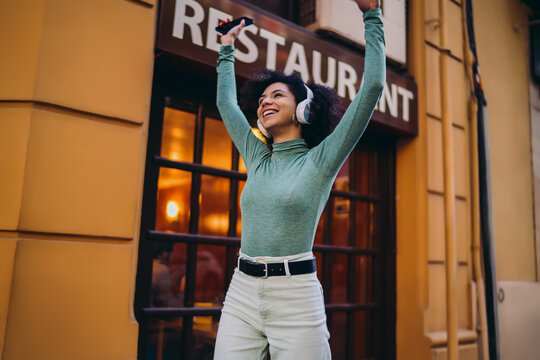 young student celebrating successful electronic online banking transaction with both arms raised high, happy Gen Z female tourist listening to music on headphones along city street