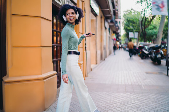 young tourist strolling along city sidewalk while listening to favorite audio track on wireless headphones, happy Gen Z user holding modern smartphone device near street store entrance