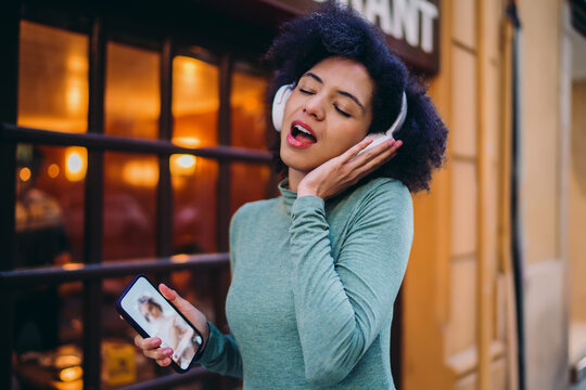 young user singing passionately while listening to favorite audio track on wireless headphones outdoors, happy Gen Z girl holding mobile smartphone device near warm restaurant display