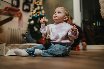Fototapeta premium A toddler girl sits on the floor in a pink floral sweater, looking up with curiosity. A cozy home scene shows a Christmas tree and gifts nearby, capturing a warm family moment.