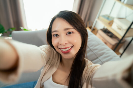 A young Asian woman is taking a selfie with her smartphone, smiling while sitting on a sofa