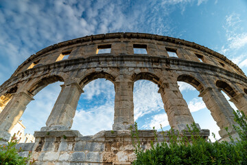 Obraz premium Pula, Croatia - August 18, 2025: Low-angle view of an ancient stone amphitheater with arched openings against a bright blue sky with scattered clouds.