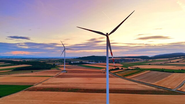 Alternative energy turbines on golden farmland at sunset. Drone view of two wind generators over yellow harvested fields under warm orange sky.