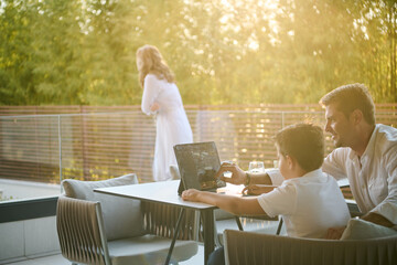 Fototapeta premium Family Spending Evening On Balcony Father Teaching Son On Laptop While Woman Looks Out