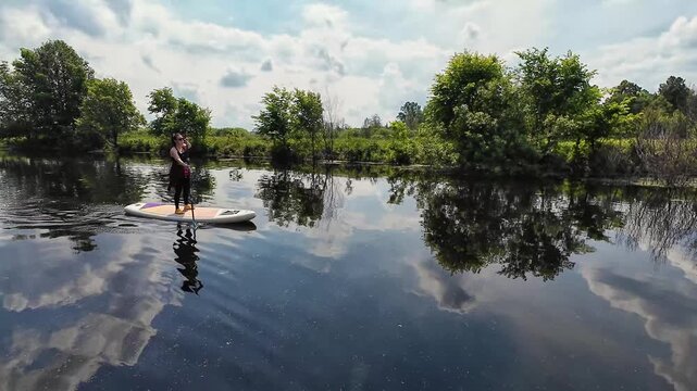 A lone person stands on a paddleboard near the left-center foreground, paddling to the right across a calm river. Soft natural light illuminates mirrored cloud reflections on the