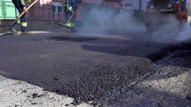 Fresh Hot Asphalt Smoking Close Up With Heavy Rollers And Laborers Teamwork. Combination compactor driving backward while construction crew uses shovels, lute tools, and wheelbarrow on roadwork site.