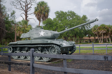 Fototapeta premium A restored historic military tank monument sits on display in a park setting with palm trees and wooden fencing. War Veterans Memorial Park In St. Petersburg, FL conveying military history, remembranc