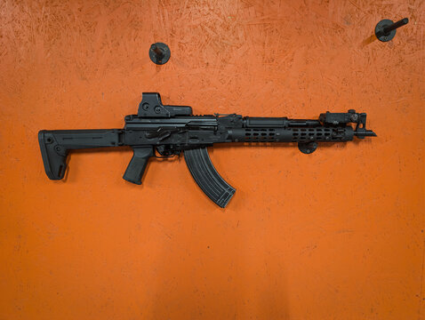 Baltic States, July 8, 2026: Modernized Soviet AKM rifle equipped with EOTech holographic sight and tactical accessories displayed on an orange wall at a shooting range.