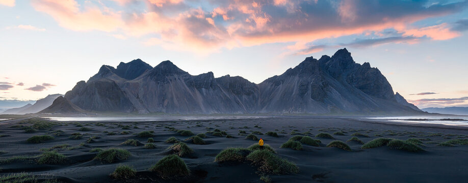 Wide panoramic view of Vestrahorn at sunset with a lone traveler standing on Iceland's black sand beach. Dramatic mountains, colorful clouds and volcanic landscape create an epic scene.