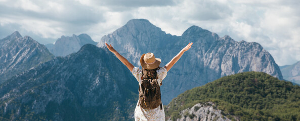A woman stands on a mountain peak, arms raised in triumph, enjoying the panoramic view of rugged mountain ranges under a cloudy sky. She is wearing a dress, backpack, and sun hat. © magic_cinema