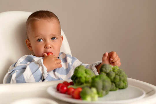 A young child sits in a high chair and enjoys eating vegetables. There are green vegetables and cherry tomatoes on a plate in front of him
