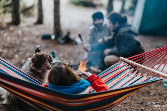 A person relaxes in a colorful striped hammock while a brown dog sits nearby. Two friends chat by a small campfire in the background, creating a cozy outdoor scene.