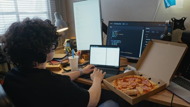 Young male programmer typing on laptop at home office desk with pizza box nearby. Footage is perfect for software development, remote work, and IT education campaigns, back view
