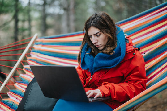 A woman works intently on a laptop while sitting in a colorful hammock outdoors. She remotely works from a forest setting, bundled in a warm red jacket and scarf.