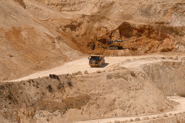 yellow caterpillar excavator digging in a large open-pit sand and stone quarry while a heavy haulage dumper truck drives along a dirt terrace road, ideal for heavy industry design layouts. © Ekaterina