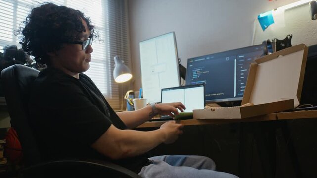 Young adult Asian man typing on laptop, picking up smartphone and eating pizza slice at desk with monitors showing code. Ideal for IT industry, remote work, and software development, low angle