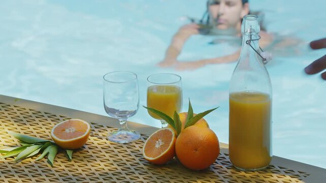 A young woman relaxes on a swimming pool while a waiter pours fresh orange juice into a glass from a bottle.