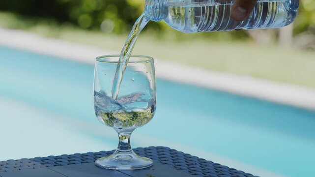 Close-up of fresh clear water being poured from a bottle into an empty glass goblet on a table by a bright blue swimming pool. 