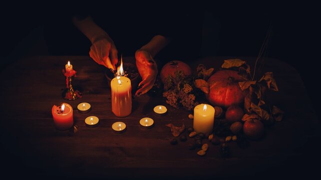 Mysterious witchy hands lighting a candles on an Samhain autumn altar next to pumpkins, herbs and magic tools in the dark.