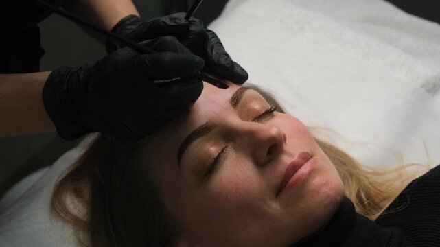 Professional cosmetologist coloring client's eyebrows with a brush during a permanent makeup procedure