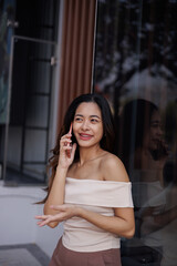 Fototapeta premium Young Asian Woman Smiling While Talking on Smartphone Outdoors Near Modern Building