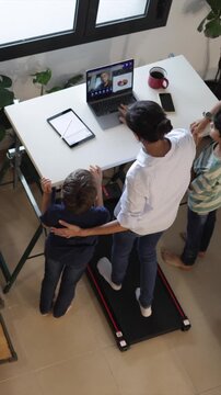 Mother juggling work from home and family life, multitasking on a treadmill desk during a video conference while her children play around her in their modern apartment